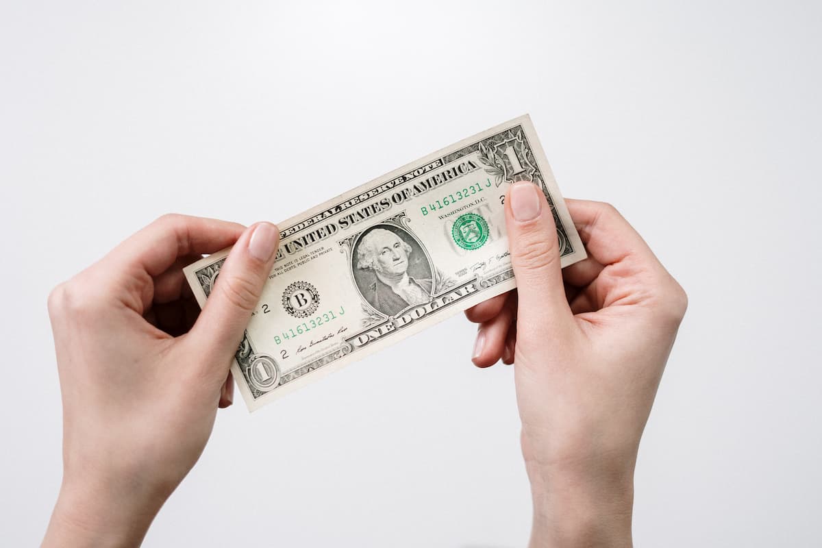 A person's hands holding a 1 US dollar bill on a white wall. 