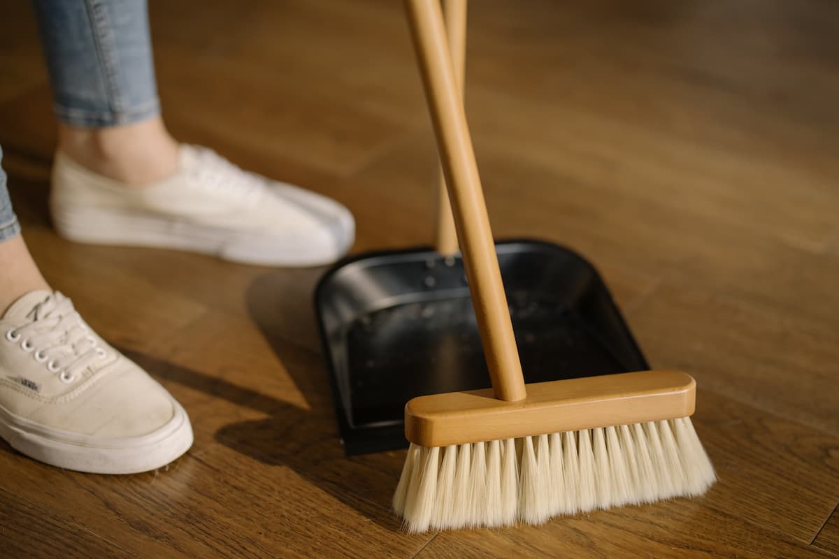 Cropped photo of a person wearing white shoes is sweeping the floor with a broom and a black dustpan.