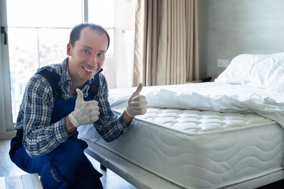 A bed bug exterminator gives two thumbs up beside a white bed.