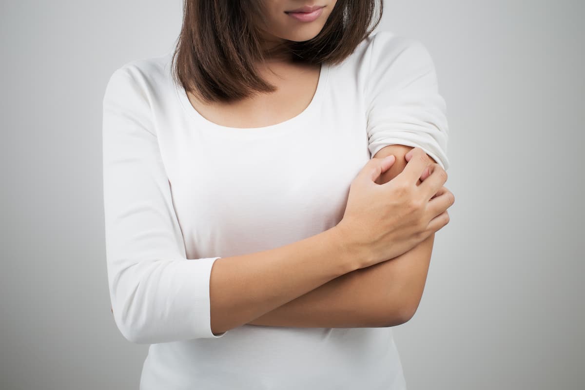 A woman wearing white long sleeves is scratching her arm.