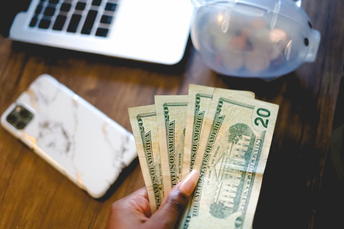 A woman's hand holding 20 US dollars against a blurred background of a phone, piggy bank, and laptop on a wooden table. 