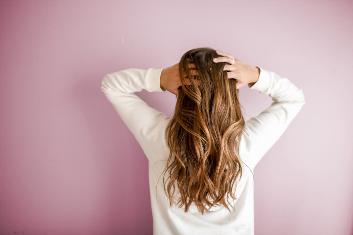 A woman in white long sleeves stands against the pink wall, holding her hair.