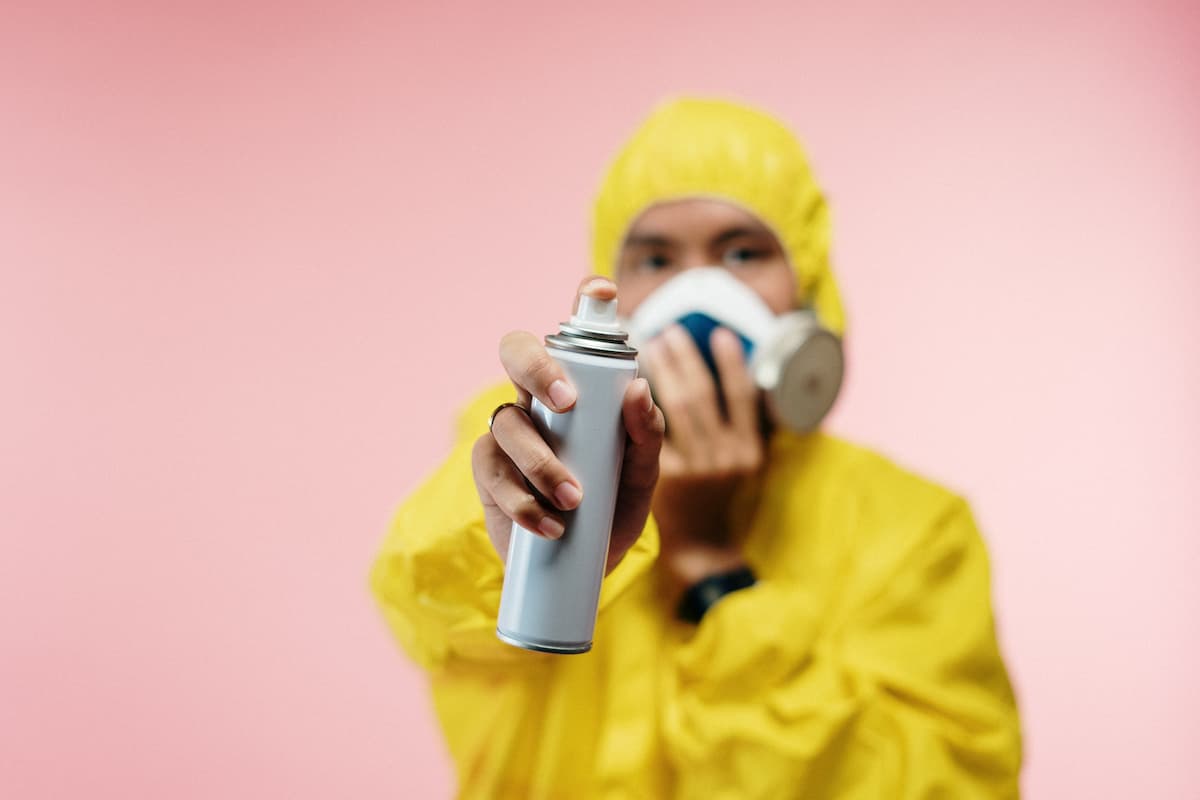 A man in a yellow coverall and mask is holding a bottle of spray on a pink background. 