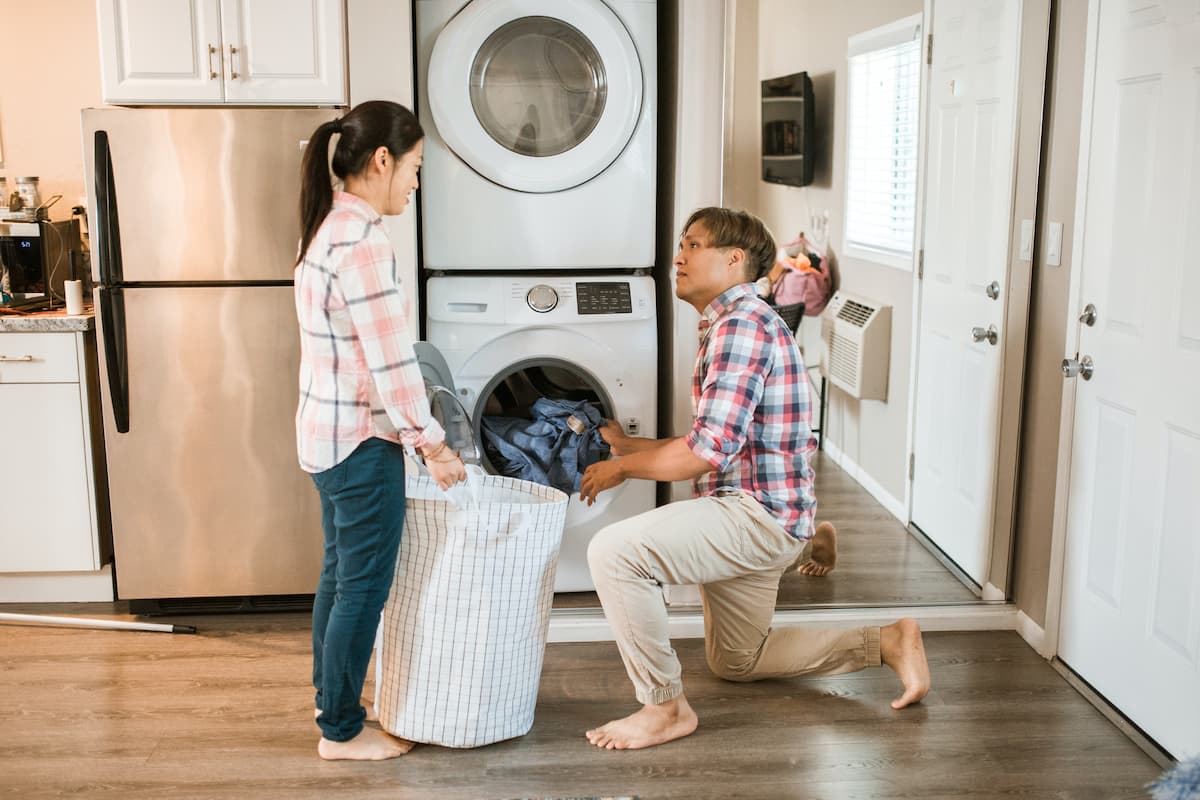 A man in plaid long sleeves is loading clothes in the washing machine and a woman wearing a plaid long sleeves is holding the laundry basket. 