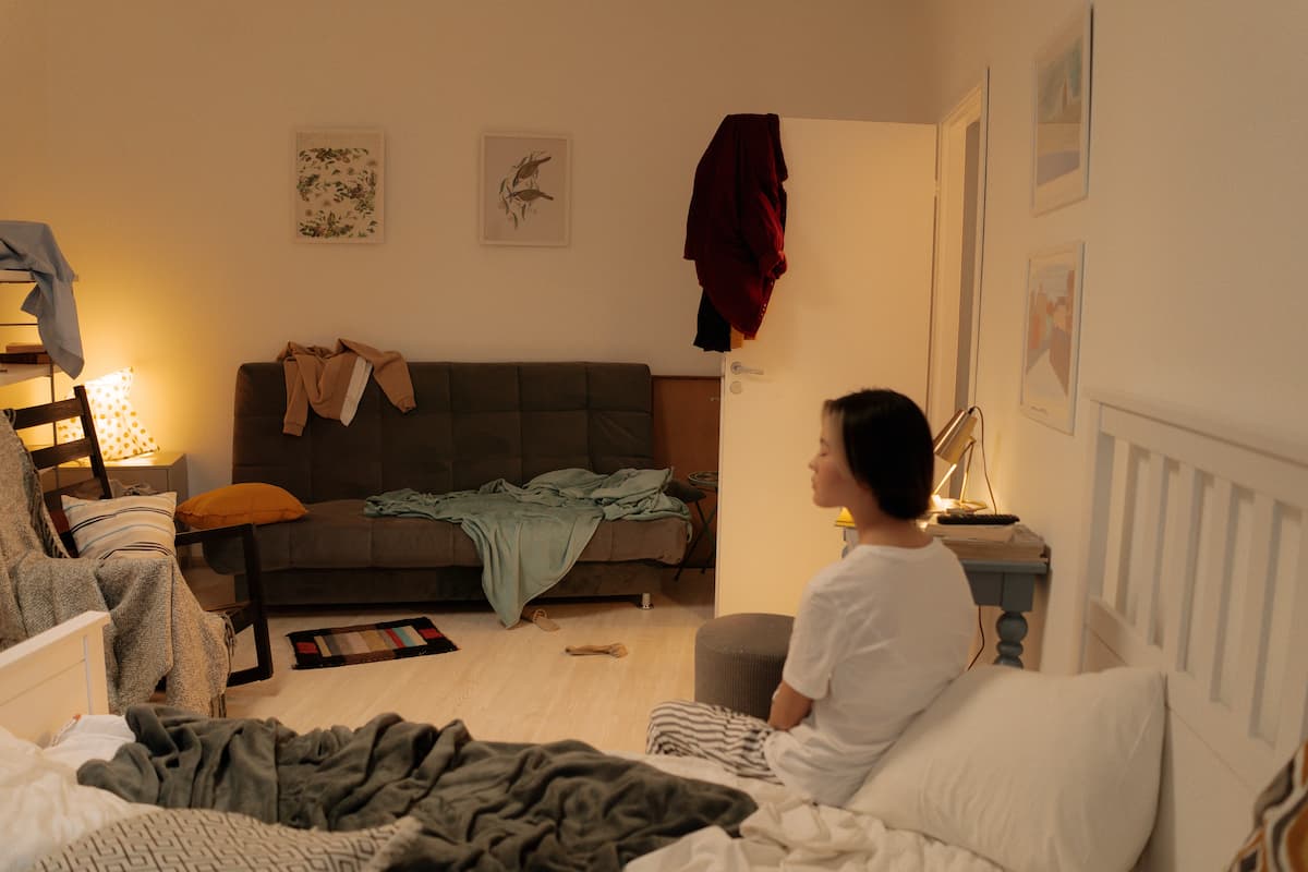 A woman in a white shirt is sitting on the bed in a messy bedroom. 