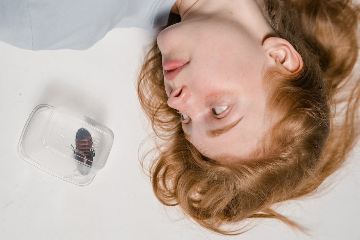 A plastic container is covering a cockroach as a terrified woman stares at it while lying down.