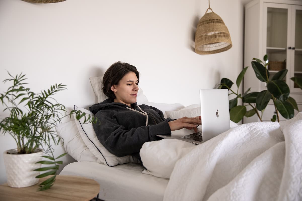 A woman wearing a hooded sweater is lying on the bed while using a laptop. 