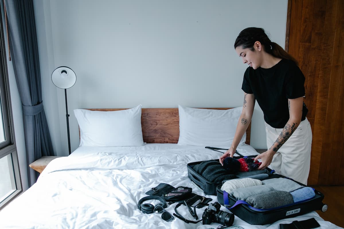 Inside a hotel, a woman in a black t-shirt packs her belongings into her luggage. 