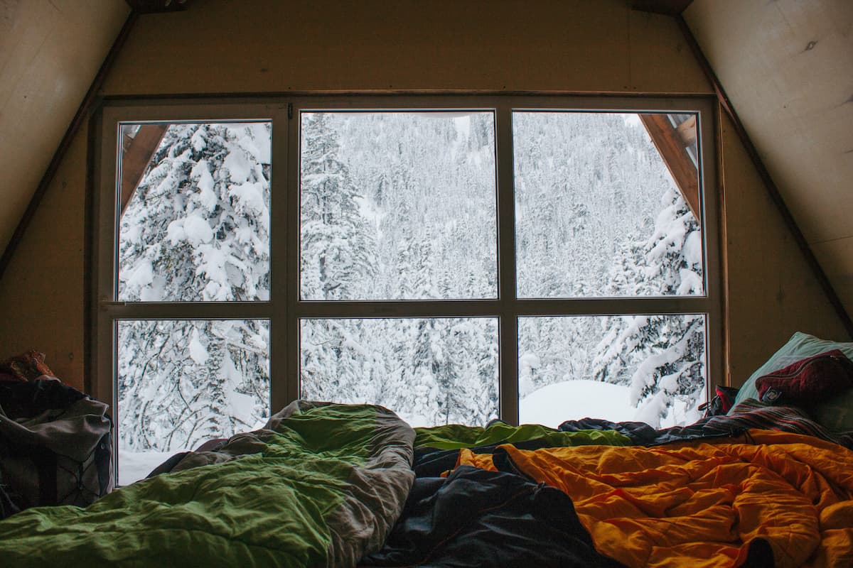 An untidy bed next to a window with a view of snow-covered trees.